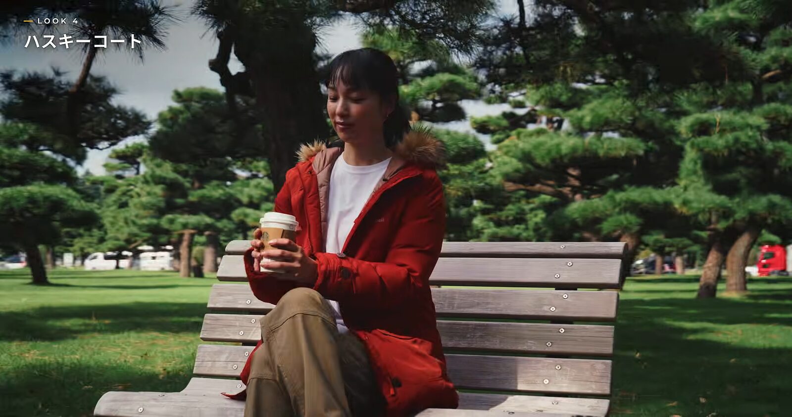 ハスキーコートにタンのパンツを合わせ、公園のベンチでコーヒーを持つ様子